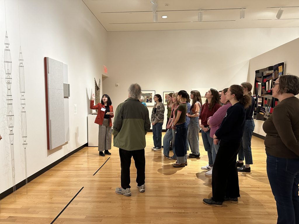 A person in a red jacket gestures to a large work of art on a wall as a group of people observe